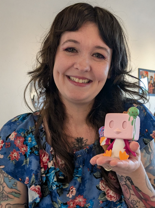 Person holding a colorful toy figure with a pink square head and a plant on top, wearing a floral blouse.